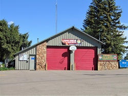 A fire station building with two red garage doors, trees, and a blue dumpster nearby.