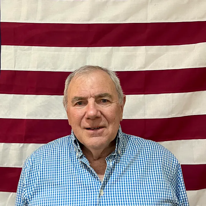 A man in a blue checkered shirt stands in front of a flag with red and white stripes.