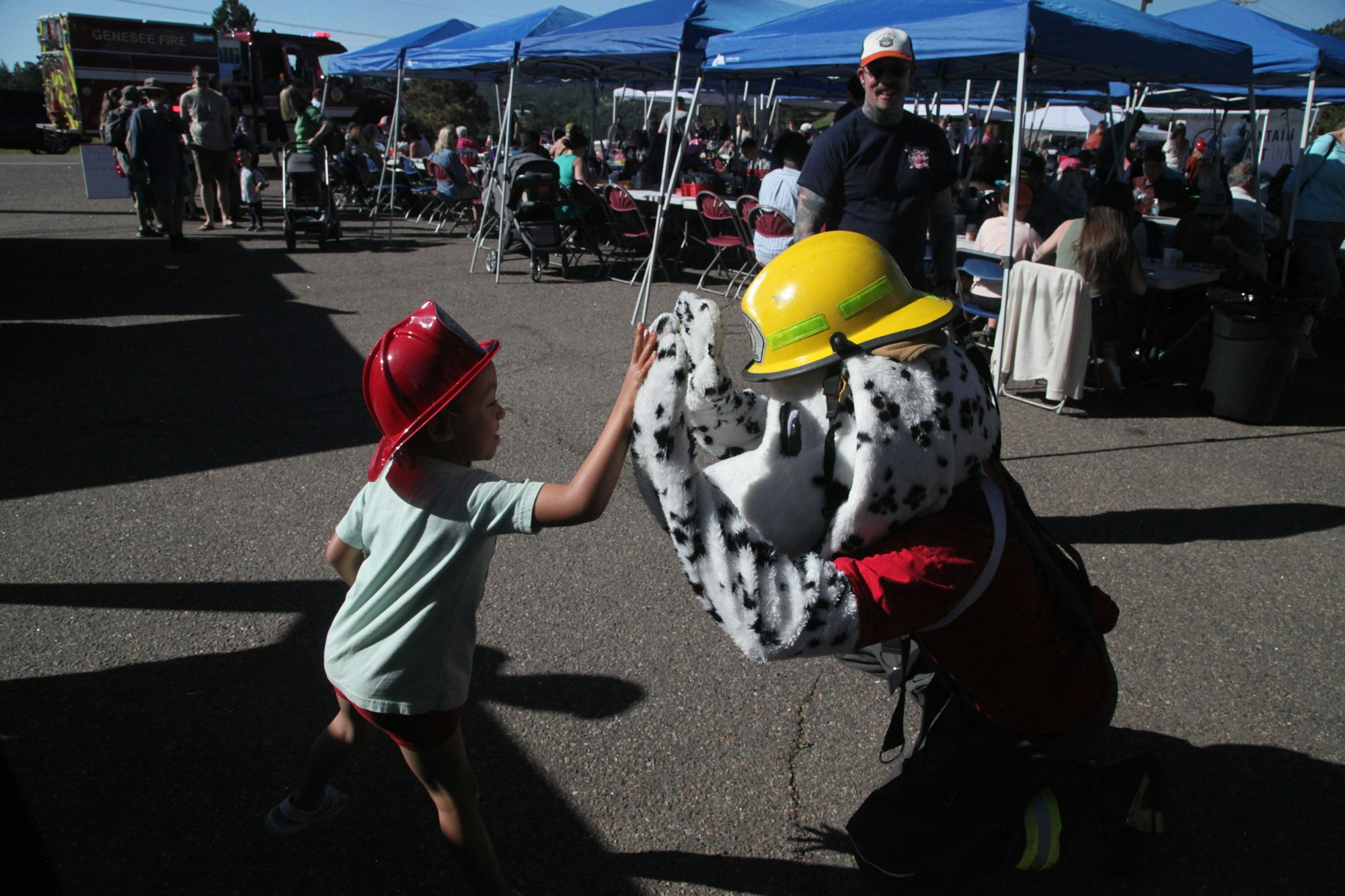 A child high-fives a person in a Dalmatian mascot costume. They're both wearing helmets at an outdoor event.