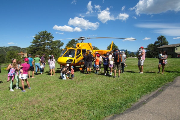A yellow helicopter on grass with a group of people gathered around, clear blue sky and trees in the background.
