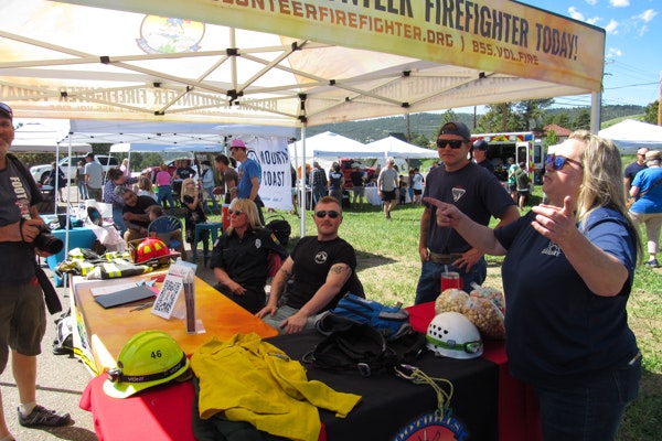 People at a volunteer firefighter recruitment booth with helmets, gear, and a banner at an outdoor event.