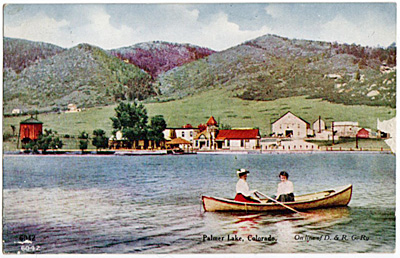 A vintage postcard of Palmer Lake, Colorado, with two people canoeing on the lake and buildings against a backdrop of hills.