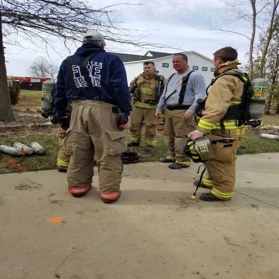 Four firefighters standing outdoors, wearing gear, with equipment nearby.
