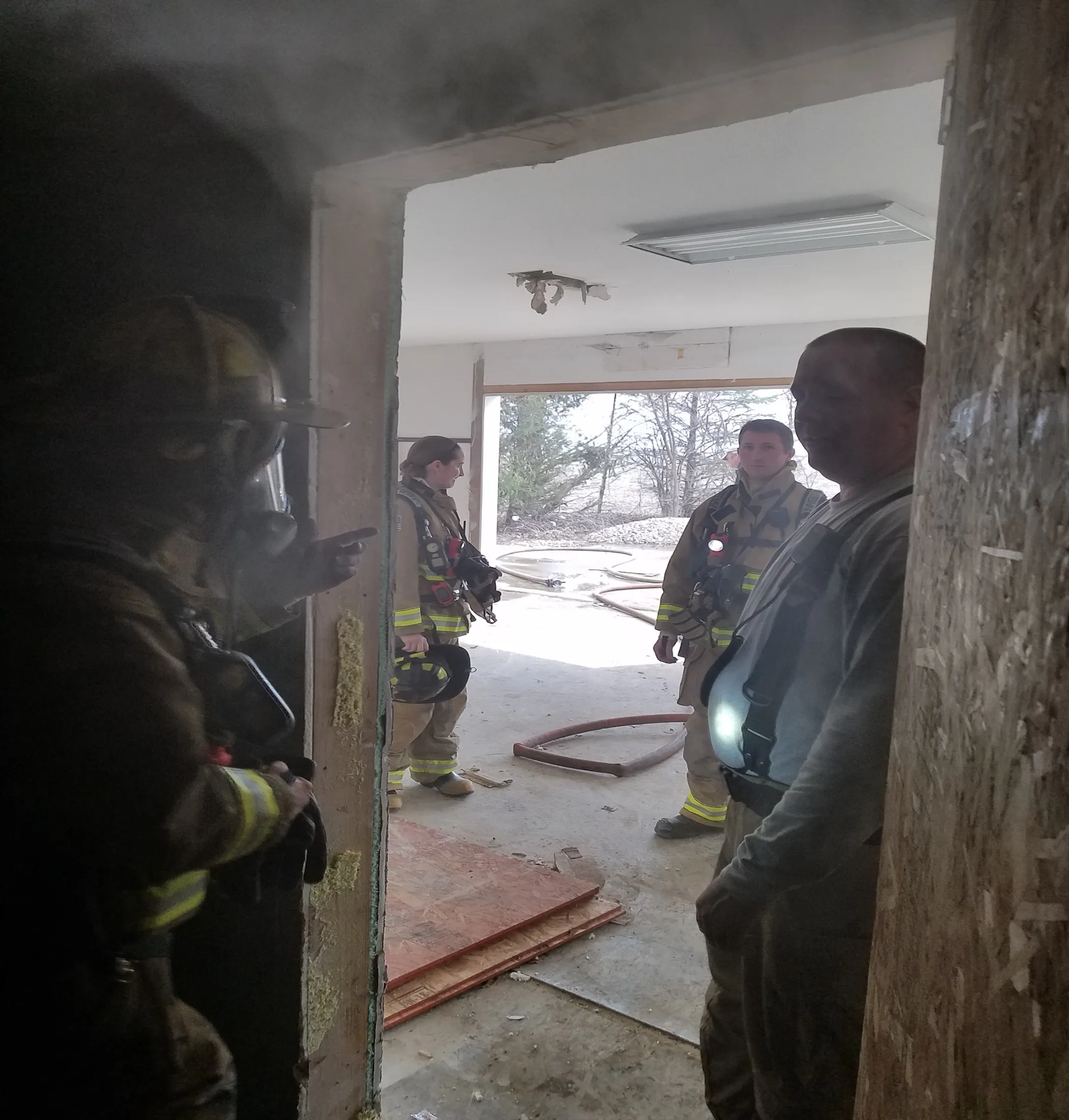 Firefighters in gear inside a smoky room with a view to the outside; one person stands in the doorway.