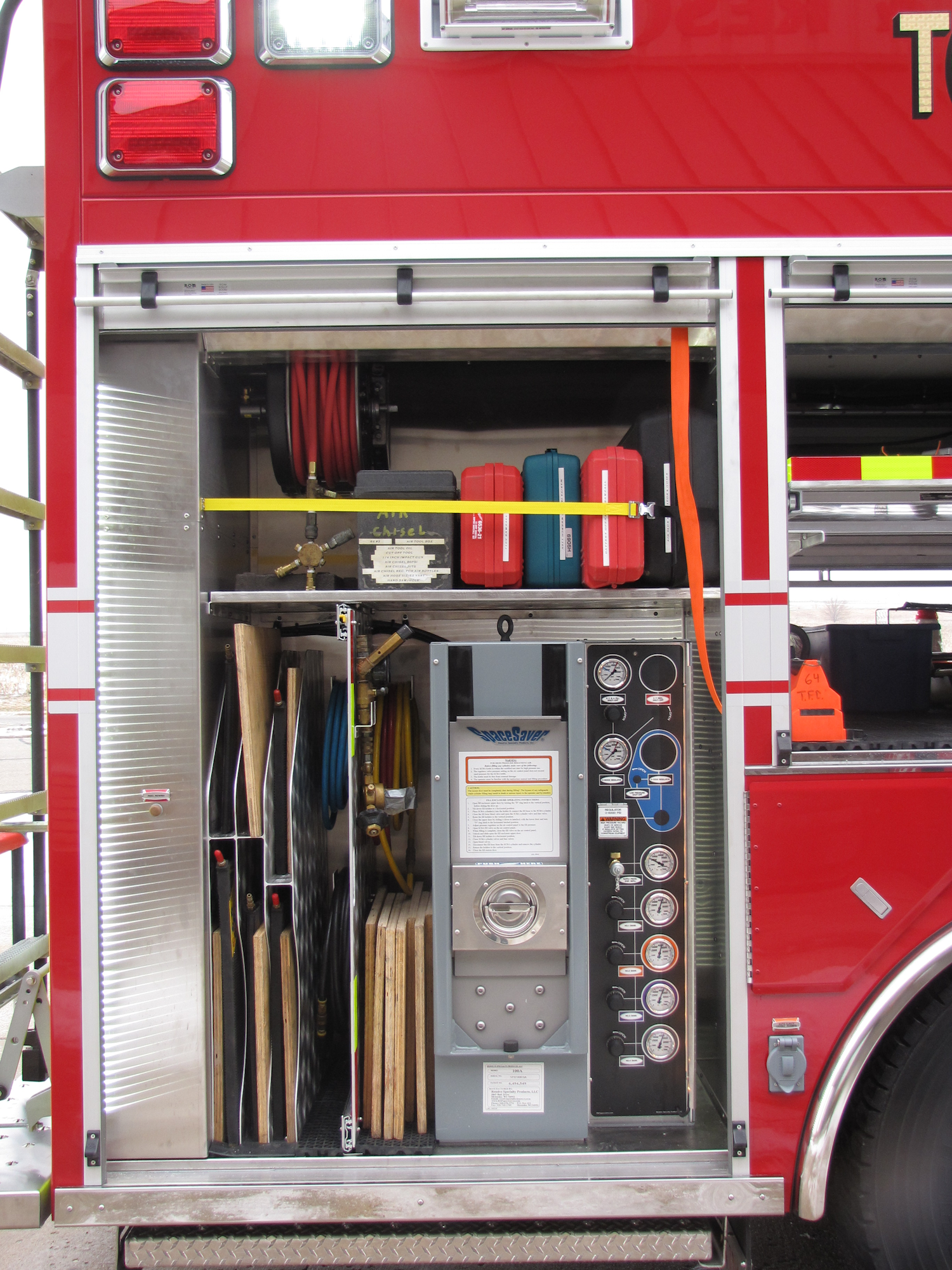 The image shows the interior of a fire truck compartment with hoses, tools, a pressure gauge panel, and safety equipment.