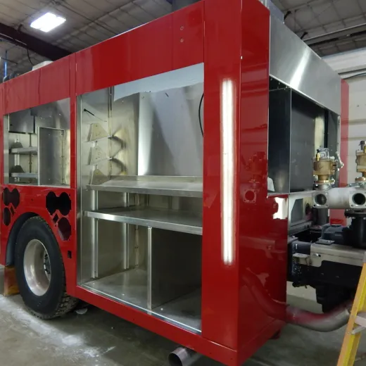 A partially assembled red fire truck chassis with storage compartments, wheels, and visible piping.