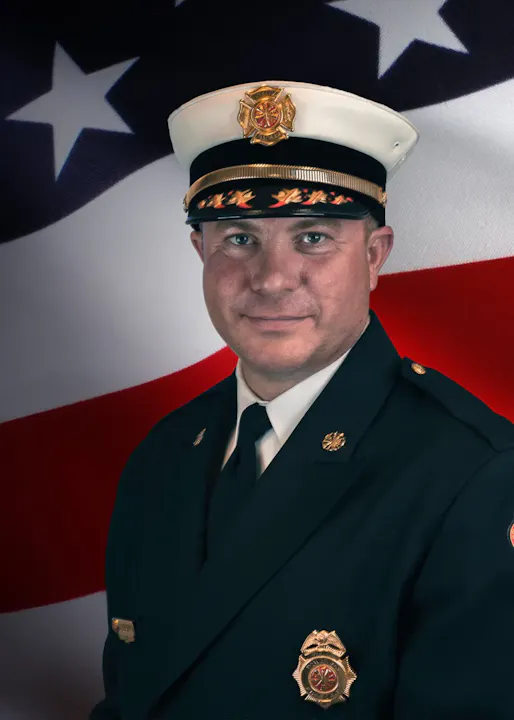 A firefighter in formal uniform with badges, standing in front of an American flag.