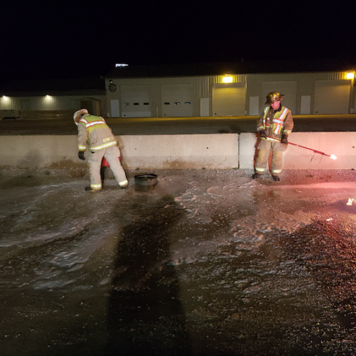 Two firefighters in gear manage a small scene at night, surrounded by foam on the ground.