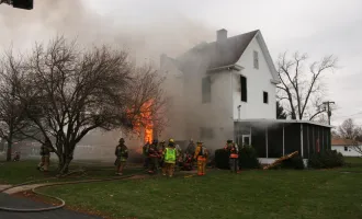 Firefighters combating a blaze in a large house, with visible flames and smoke billowing.