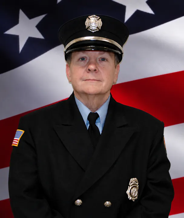 A person in a police or firefighter uniform posed in front of an American flag background.