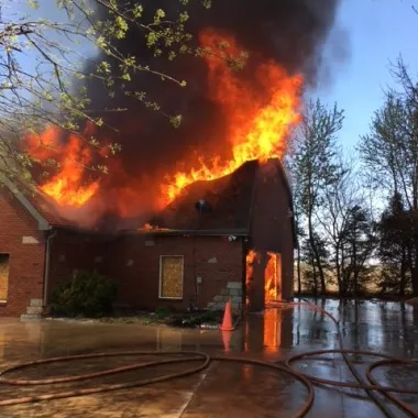 A brick house is engulfed in flames, with heavy smoke and nearby trees. Fire hoses and water are on the ground.