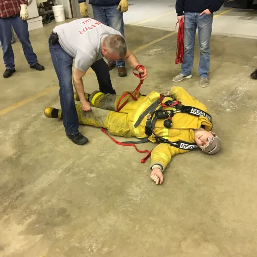 A person is lying on the floor in a yellow suit, possibly for a safety demonstration, while someone assists with a red strap.