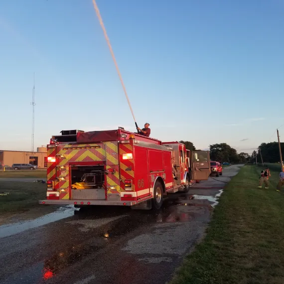 A firefighter on a fire truck sprays water from a hose, with emergency vehicles and personnel nearby.