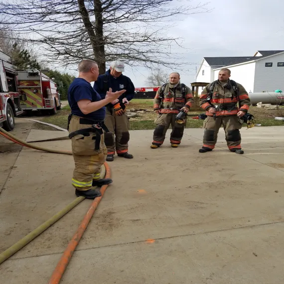 Four firefighters in gear having a discussion beside a fire truck outside.
