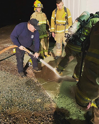 Firefighters during training or an operation, with one handling a hose spraying water on the ground.