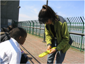 Adult showing a child something yellow, outdoors near a green fence, sunny day.
