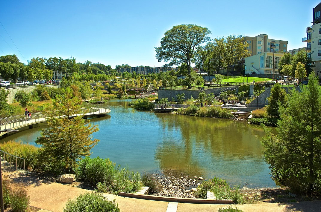 Urban park with a pond, bridge, walkways, greenery, and buildings in the background under a clear blue sky.