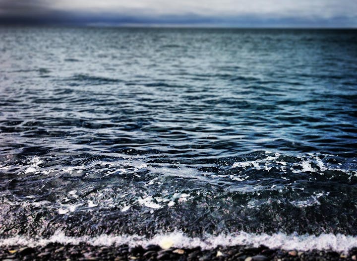 A close-up of a gentle wave washing onto a pebbly shore with overcast skies above.