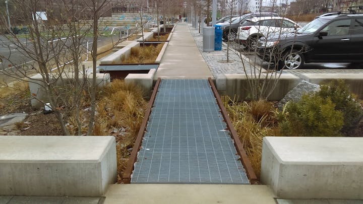 Urban walkway with benches, planters, and a grated path; cars parked beside.