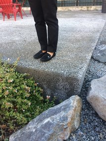 Person standing on a concrete edge with a gravel ground, rocks, plants around, and a red chair in the background.