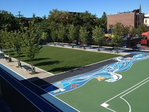 An outdoor basketball court with artistic patterns, surrounded by trees and buildings under a clear sky.