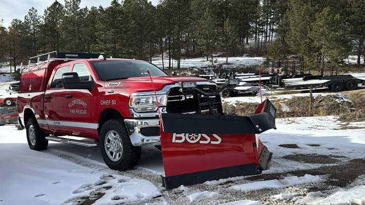 Red fire rescue truck with a snow plow attachment, parked on a snowy ground in a forested area.
