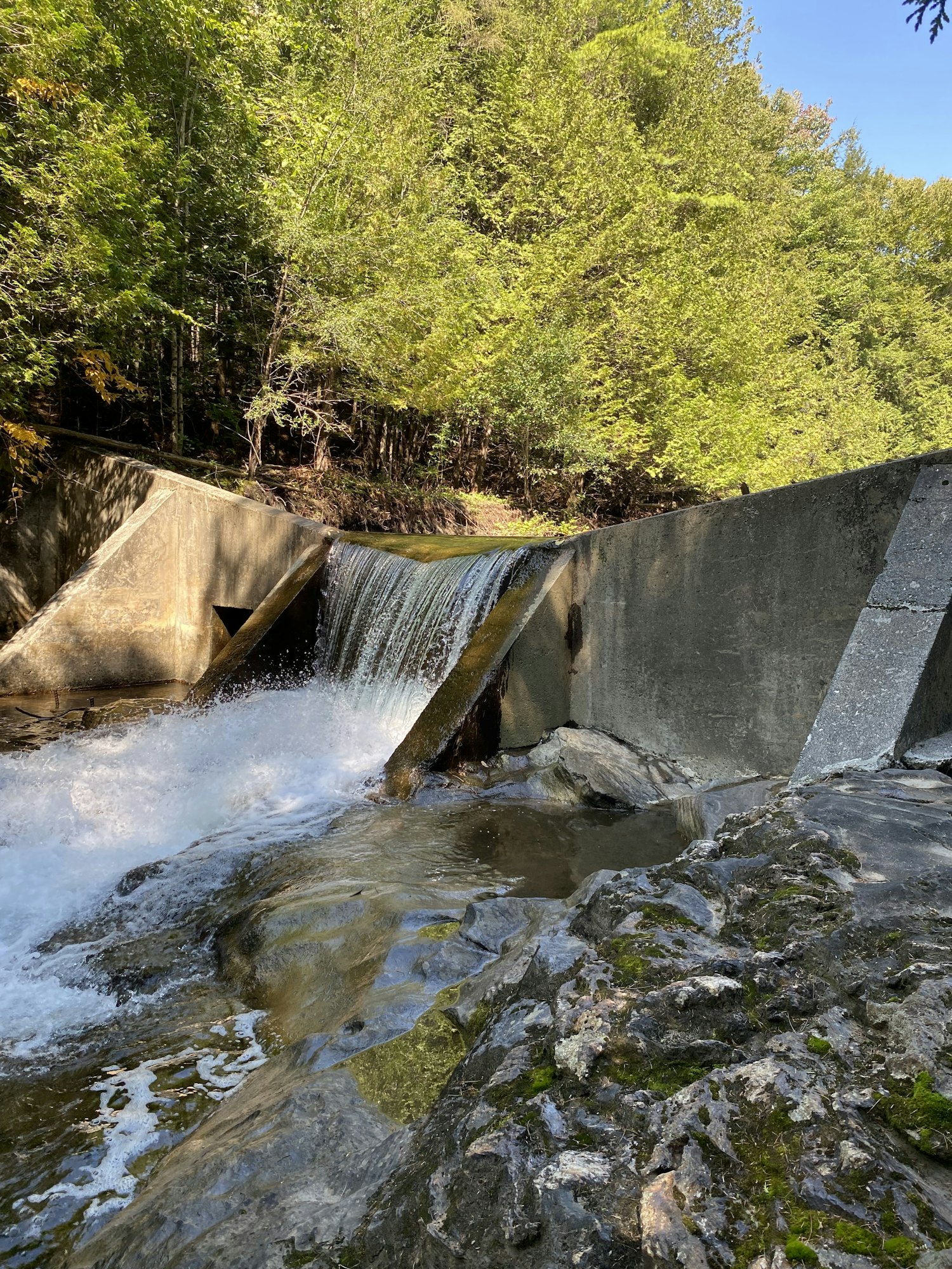 A cascading waterfall flows over a concrete barrier, surrounded by lush greenery and rocky terrain under a clear blue sky.