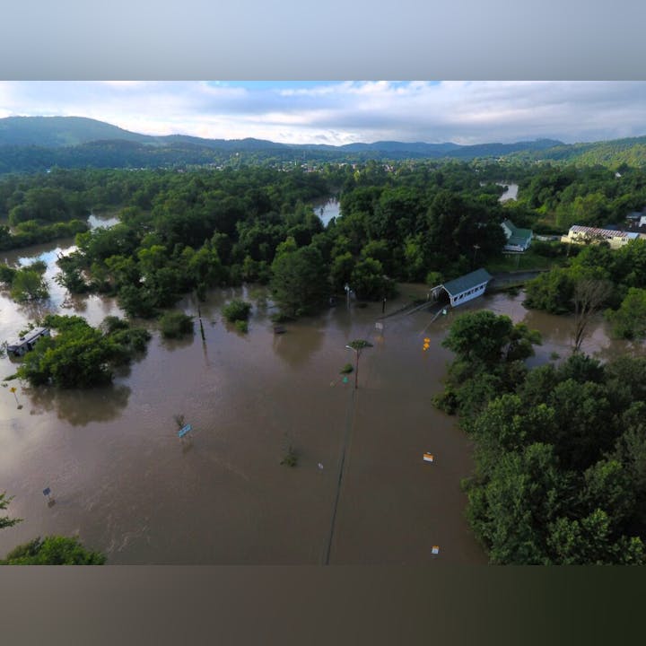 Aerial view of a flooded area with submerged roads, trees, and a partially visible structure, surrounded by lush greenery and hills.