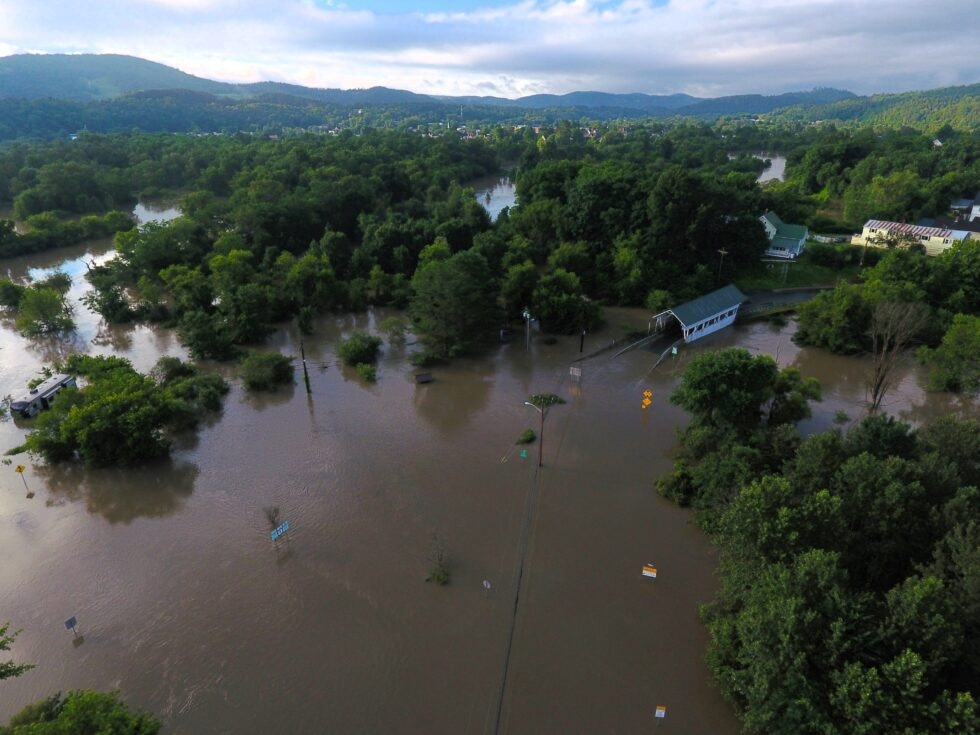 Aerial view of a flooded area with submerged roads, trees, and a partially visible structure, surrounded by lush greenery and hills.