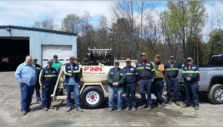 A group of workers standing in front of a Finn T60 Hydro Seeder machine next to a building and a truck in an outdoor setting.