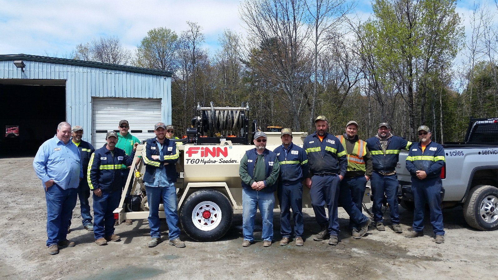 A group of workers standing in front of a Finn T60 Hydro Seeder machine next to a building and a truck in an outdoor setting.
