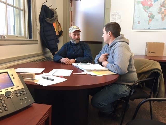 Two people sit at a round table in an office, engaged in conversation. Papers and a phone are on the table. Coats hang nearby.