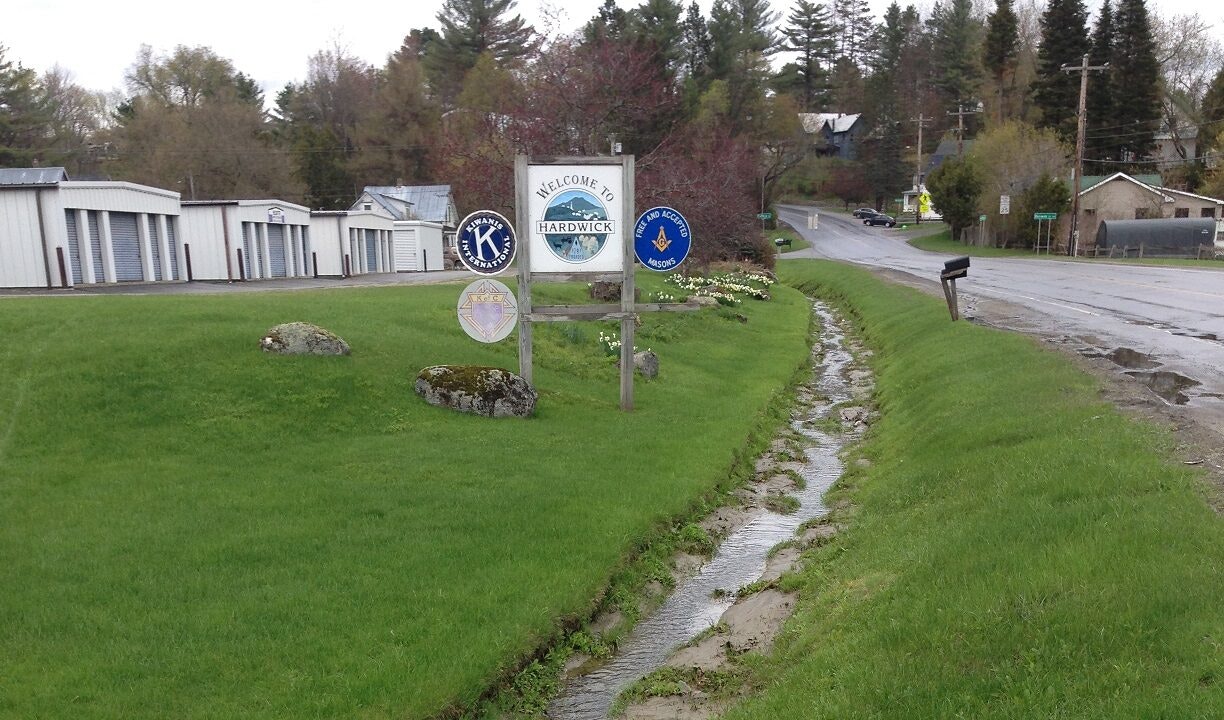 A roadside sign welcomes to Hardwick. It features logos of Kiwanis International and Freemasons. Storage units and a small creek nearby.