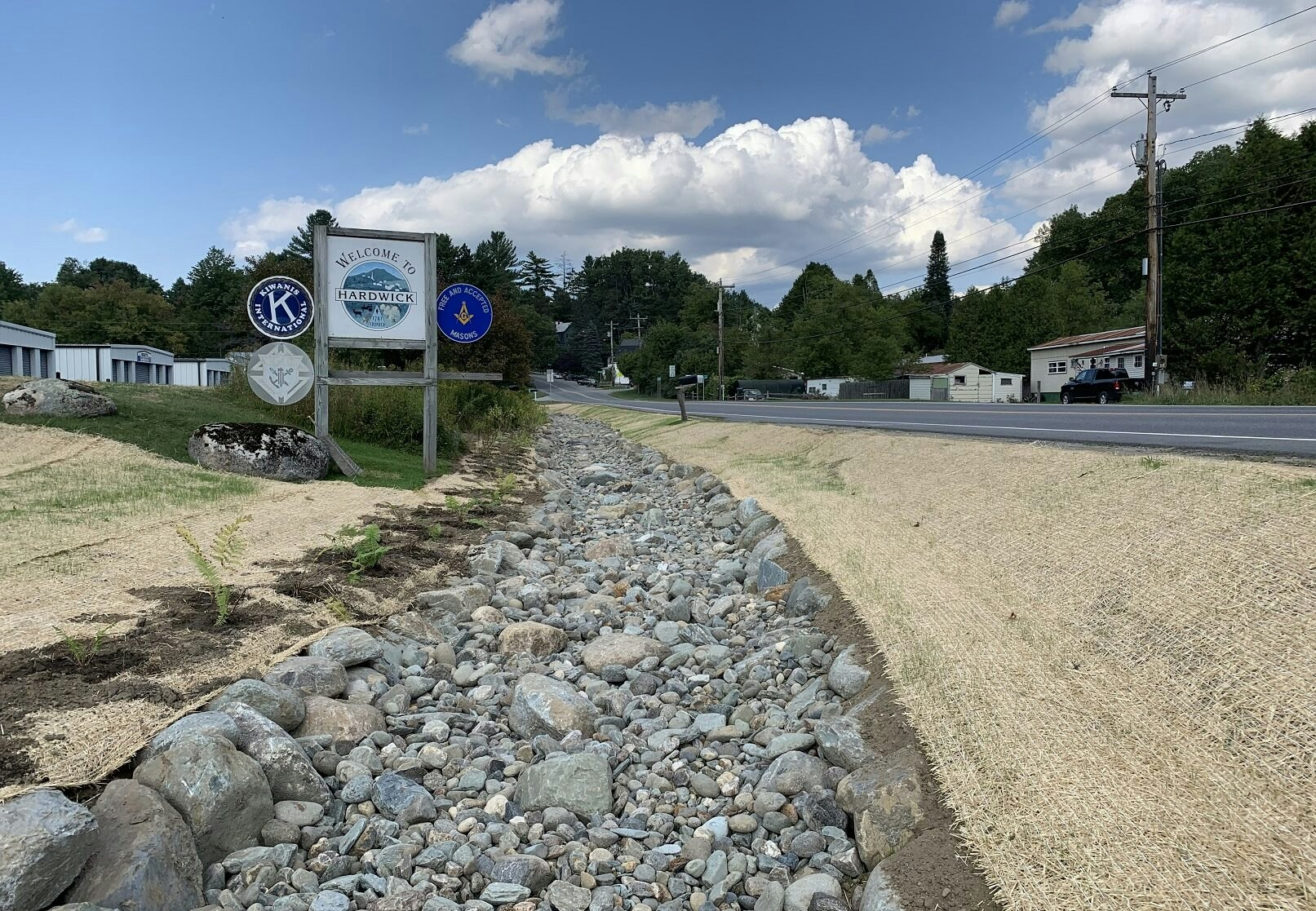 Welcome sign for Hardwick with community logos, next to a road and rocky drainage ditch under a cloudy sky.