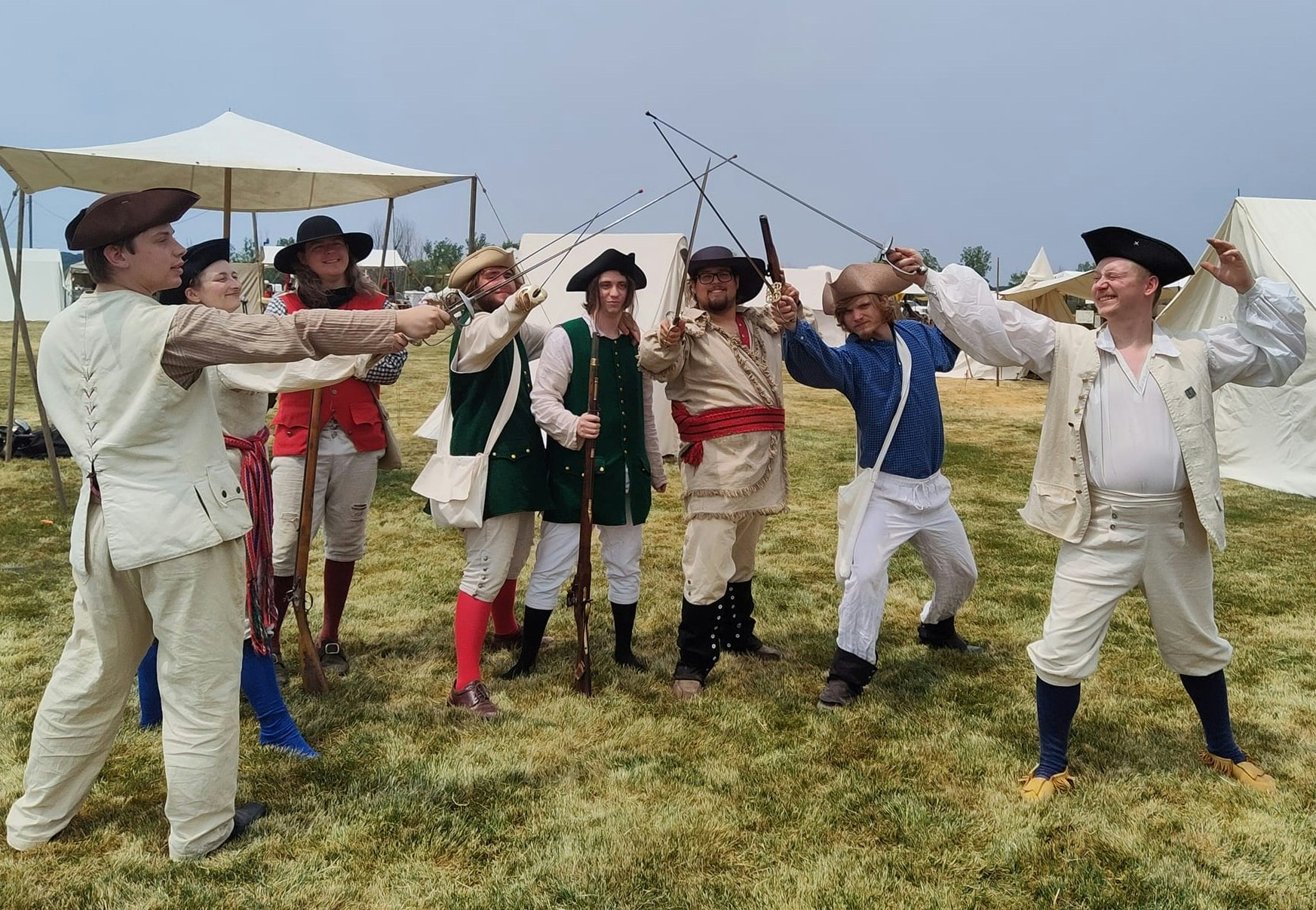 A group of people in historical costumes, posing with swords and rifles, likely at a reenactment event outdoors.