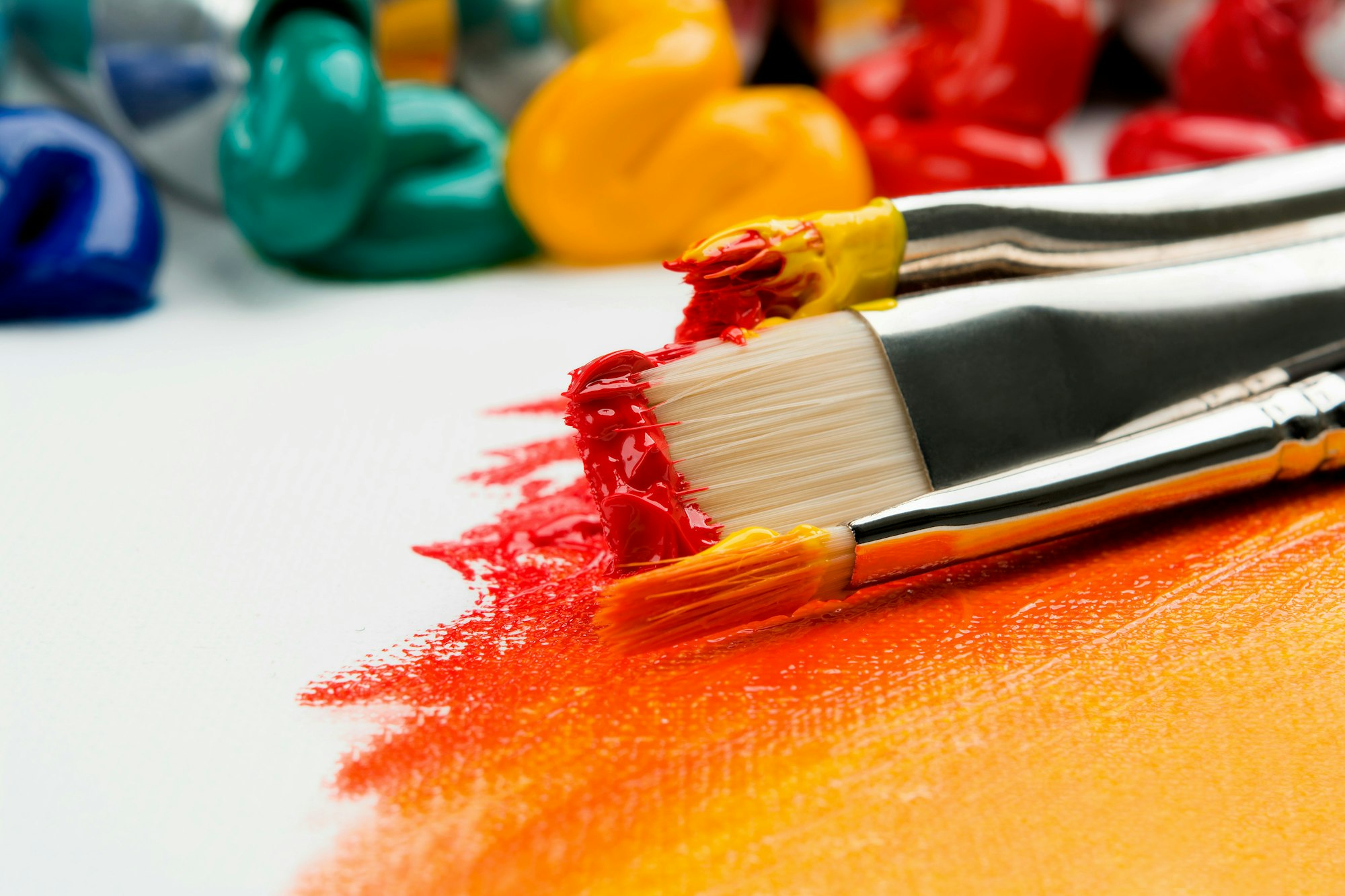 Paintbrushes with red and yellow paint on a canvas, with colorful paint blobs in the background.