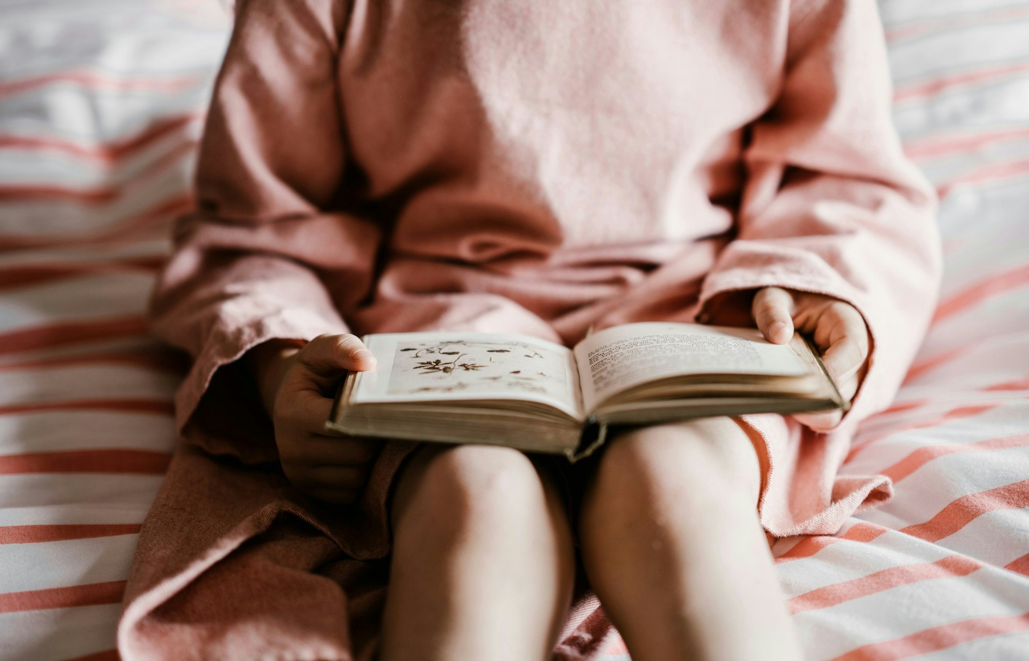 A person in a pink robe reading a book on a striped bed.