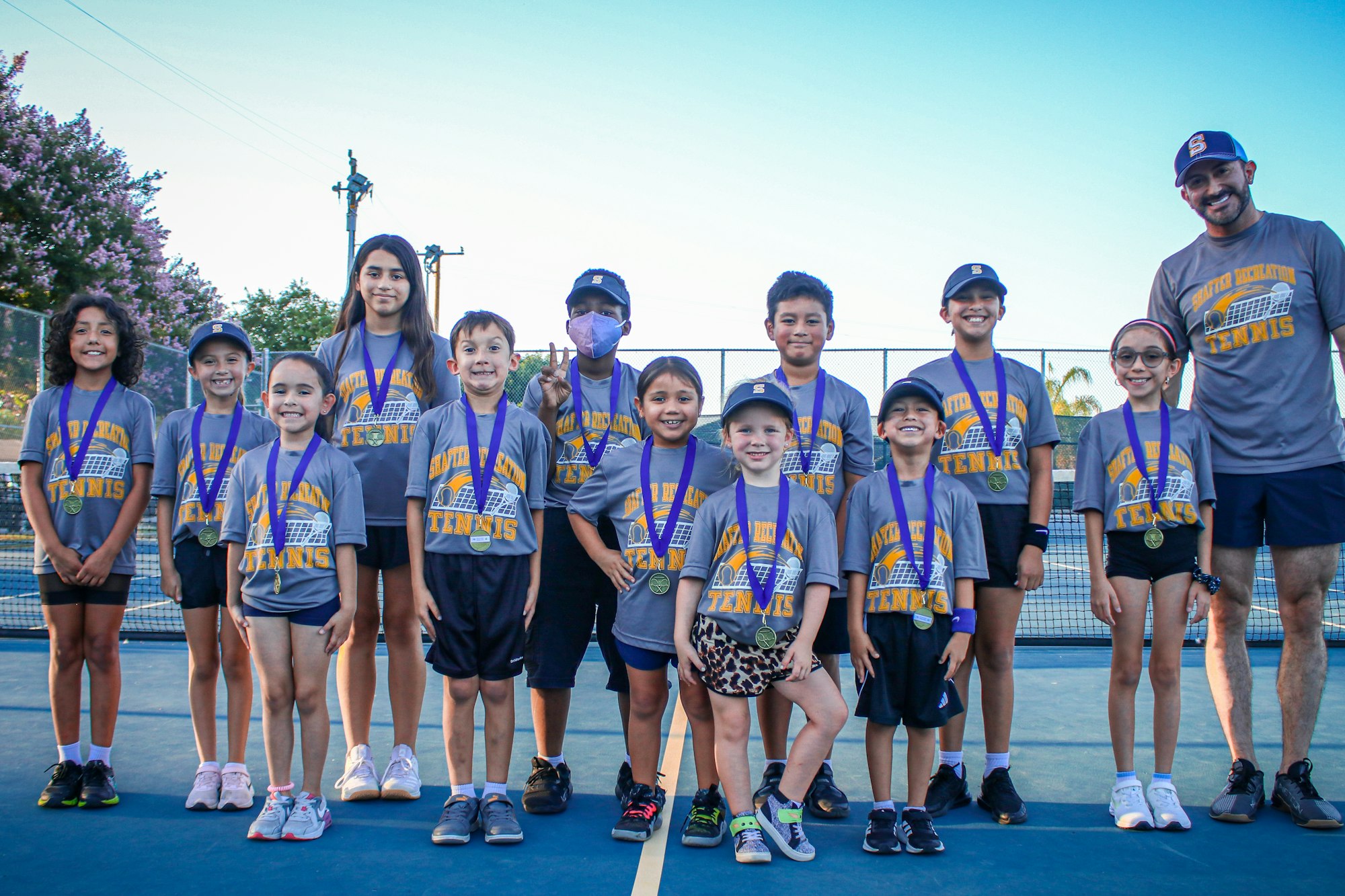 A happy group of kids with medals and an adult on a tennis court, suggesting a team photo after a sports competition.