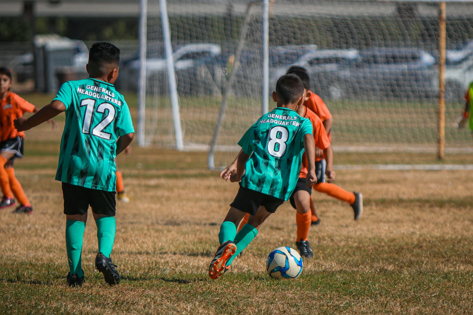 Kids playing soccer, wearing green and orange jerseys, on a grass field.