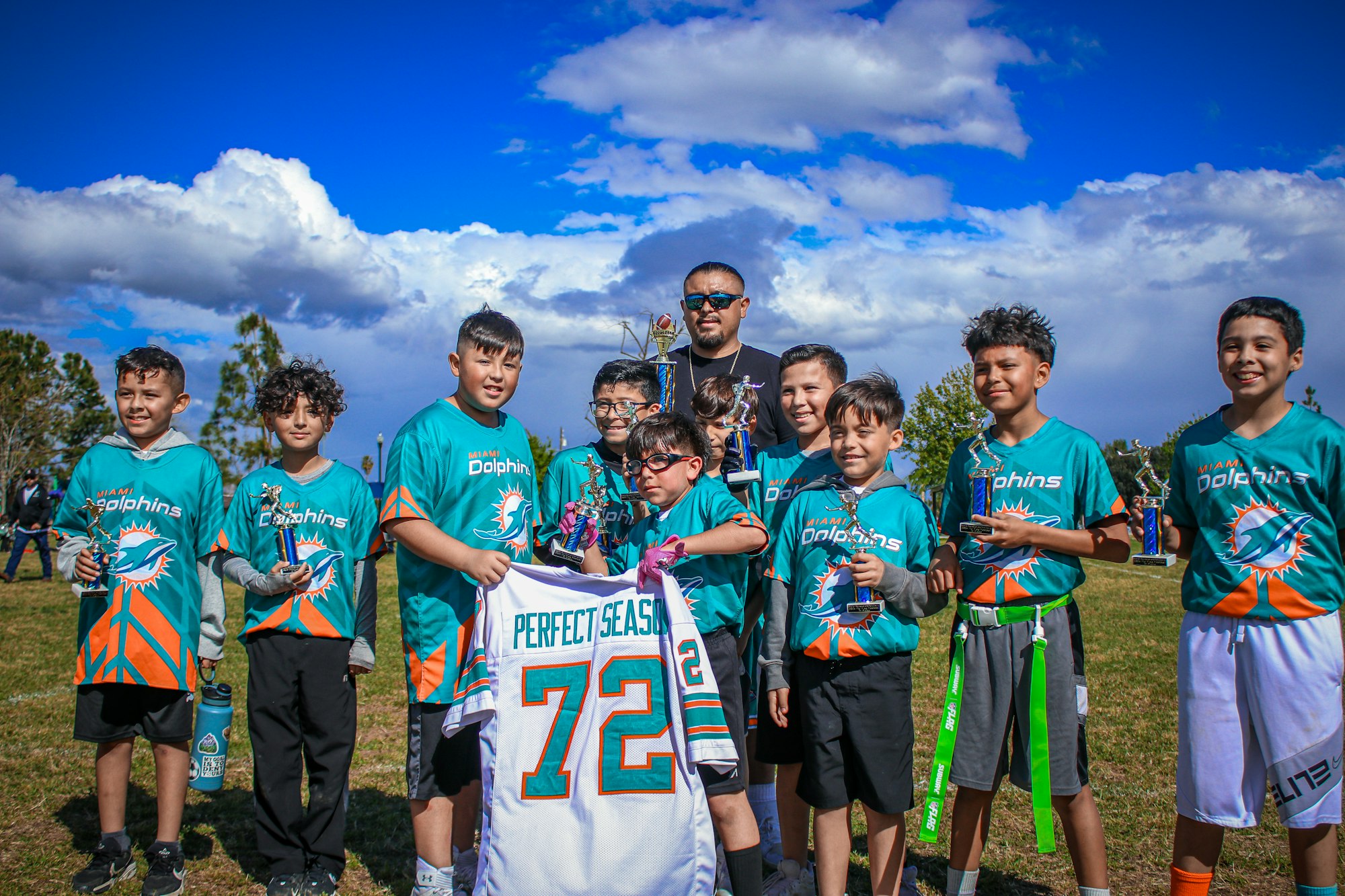 A youth sports team in Miami Dolphins attire with trophies and an adult coach.