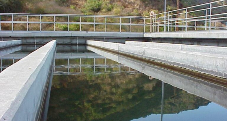The image shows a water treatment facility with concrete channels and calm water reflecting the surrounding landscape.