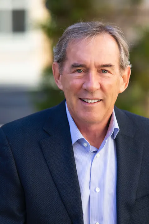 The image features a smiling man in a blazer and button-up shirt, outdoors with a blurred background.
