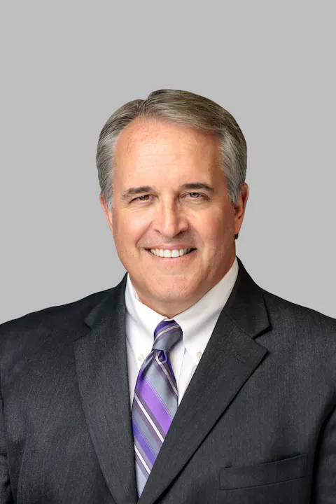A man in a suit and tie is smiling, with short gray hair and a light gray background.