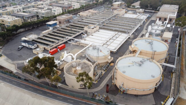 The image shows a wastewater treatment facility with large tanks, processing areas, and surrounding urban development.