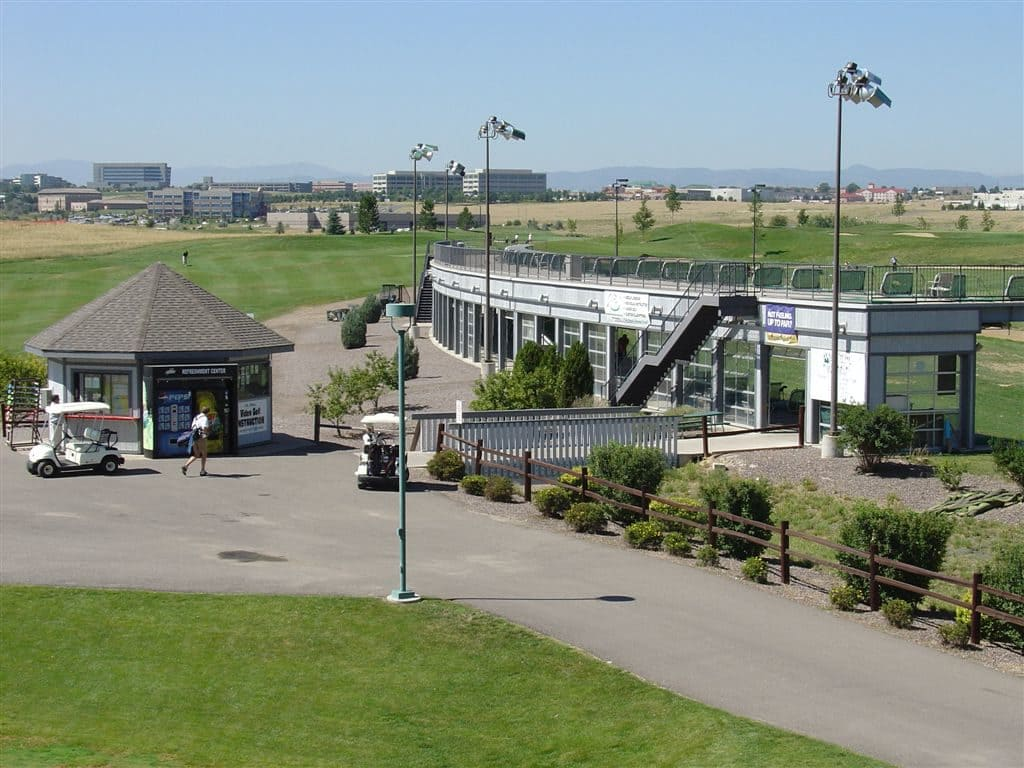 A golf driving range with a two-story building, golf carts, a path, and a person walking. Buildings and fields in the background.