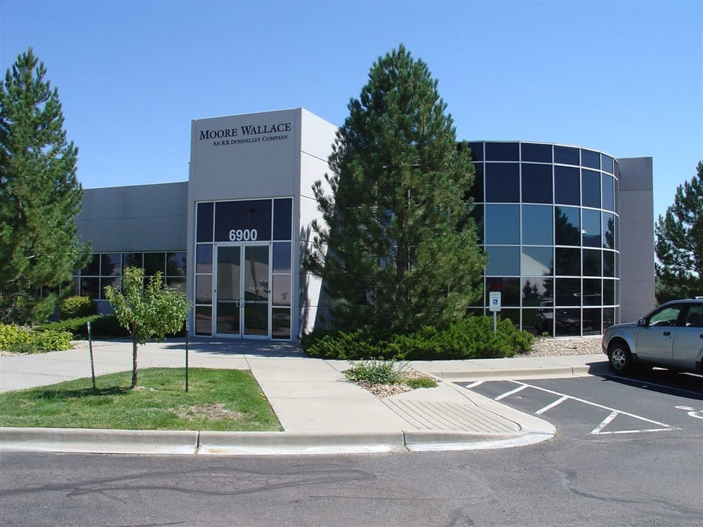 Commercial building with "Moore Wallace" sign, surrounded by trees. The address is 6900 with a parking area in front, under a clear sky.