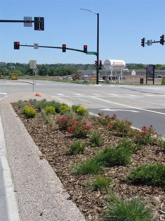 Intersection with traffic lights, landscaped median with flowers, and a visible water tower in the background.