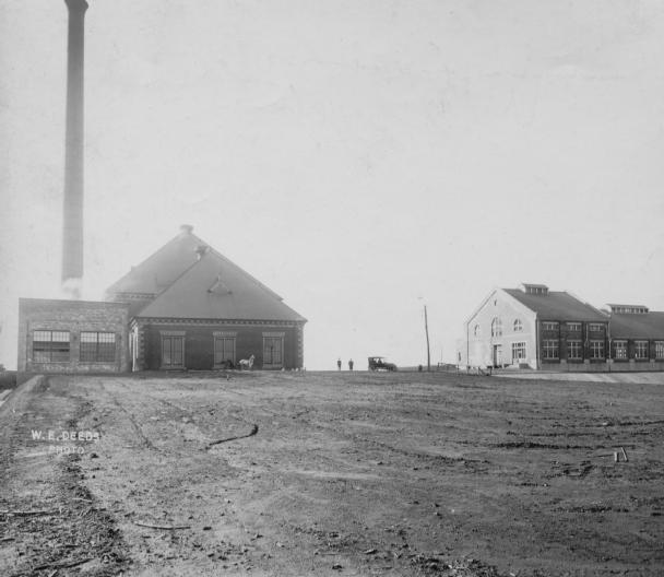 Historic industrial buildings, tall chimney, barren foreground, overcast sky.