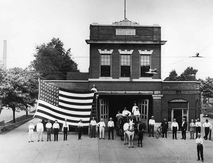 Group of people holding a large American flag in front of a vintage building with "BUS LINE" sign.