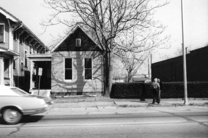 A vintage black-and-white photo featuring a house, a moving car, and two people standing by a hedge.